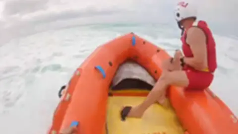 An RNLI inshore rescue boat. There is a man wearing a red life jacket and red shorts. In the background are light blue waves.