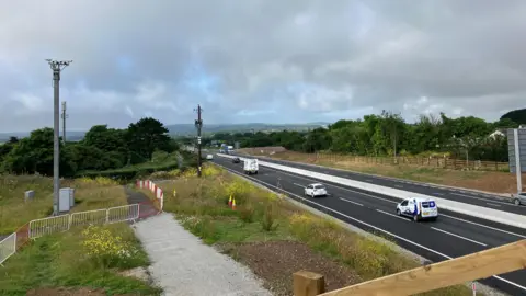 Cars and vans travel along the A30 in Cornwall on a cloudy day. Grass verges and fields are on either side of the dual carriageway.
