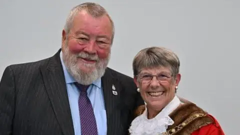 Peterborough City Council Judy Fox on the right, wearing a red ceremonial robe and a golden necklace, with John Fox standing on the left, wearing a grey striped suit with a blue shirt and purple tie.