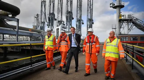 Getty Images A man in a dark grey suits walks outside on an industrial site with four men in orange high-vis clothing and hard hats. Behind them is port apparatus and machinery on a dock