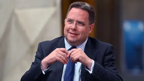 Getty Images Craig Hoy, a man with short dark hair and glasses, photographed in a medium close-up. His hands are touching in front of his chest. He is wearing a dark suit, chequered blue and white shirt and navy blue tie. 