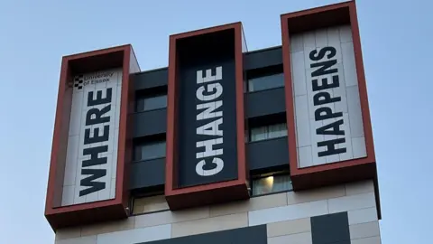 Large lettering at the top of a building that reads "Where change happens". Each of the three words is in a separate section of the facade, with the words written out vertically, starting with the first letter on its side at the bottom.