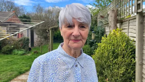 A woman wearing a blue and white blouse stands in garden. Trees, bushes and a fence can be seen in the background.