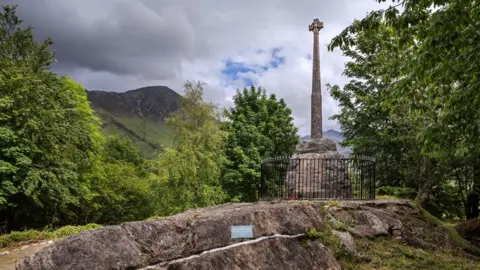 Getty Images The memorial has a stone cross atop a cairn. There is a plaque and the memorial is surrounded by a black metal railings. The site is surrounded by trees and there are hills in the background.