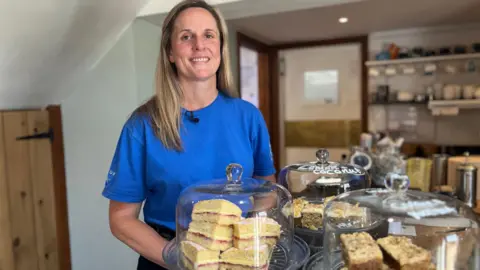 Charlotte Carter stands by a counter in her cafe, in front of her are homemade cakes. She has long blonde hair and wears a blue Hilltop Farm branded T-shirt.