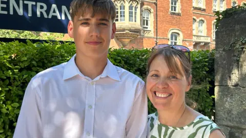A woman wearing a green and white patterned dress is standing next to a boy with a white shirt. They are next to a hedge and a stone wall. Further behind them is a red brick building with stone window surrounds and elaborate bay windows.