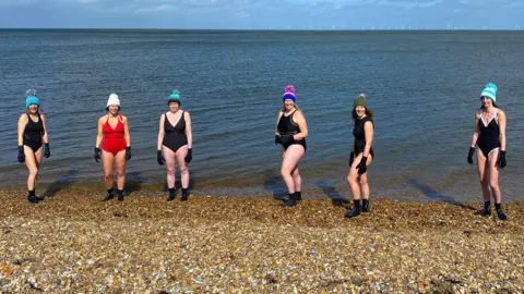 Getty Images Six mature women standing on a pebbly beach by the sea. They are wearing swimming costumes, woollen hats, black gloves and ankle-high water shoes.