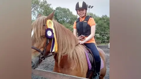 Family handout Caris Graham is sitting on a horse which has two rosettes hanging from its bridle. The horse is brown with a white patch along its nose. Ms Graham is holding a mobile phone in one hand and the bridle with the other. She is wearing an orange t-shirt and a black protective vest and riding helmet.