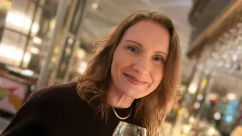Family Handout A woman in a burgundy top looking at the camera smiling. She is holding a wine glass and behind her is a out of a focus pub.