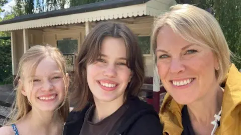 A woman and two girls smile together outdoors. Behind them is a vintage bus shelter.