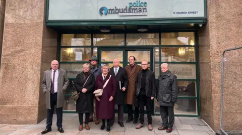 Six men and two women stand outside the glass doors of the Police Ombudsman office building in Belfast. All are wearing coats and one of the men is wearing a hat