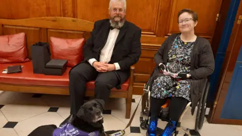 Cheryl Alexander A woman in a wheelchair sits next to a man in a tuxedo in a fancy venue. A black Labrador lays on the floor beside them.