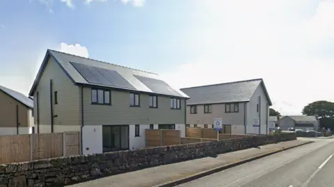 Google A street view of semi-detached homes lining a road. They have solar panels on the roof and green cladding, with wooden fences separating the homes. A number of for sale signs can be seen in the garden of the homes. 