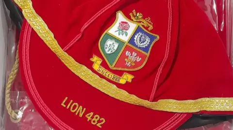 A red cap, adorned wit the crest of the british and irish lions rugby team. The cap has gold trim, and the words Lion and number 482 is written on it.