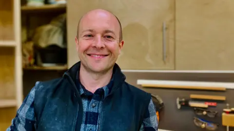Furniture maker Nick James is in his furniture making workshop in Newcastle.  He is looking at the camera and smiling and wearing his casual clothing. Behind him you can see his wood-working tools on the wall.           