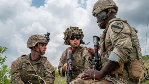 Three US soldiers in a field, wearing helmets and holding weapons and tactical gear