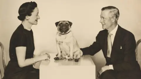 Royal Collection Enterprises Ltd 2025 | Royal Collection Trust A black and white photograph showing a man and woman sat opposite each other on two chairs smiling. A pug dog is sat on a table in the middle of them looking at the camera. 
