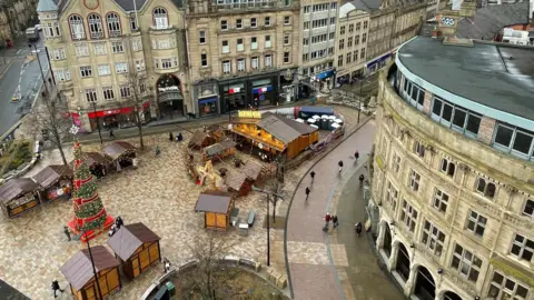 BBC/Simon Thake A view high up looking down at a Christmas Market with a decorated Christmas tree and wooden log cabins