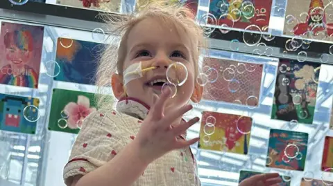 Supplied Avarni Mae surrounded by bubbles. She is looking up with one of her hands in the air. She is wearing a beige shirt with pink hearts on it. She has messy, blonde hair which is short and has a yellow tube in her nose.