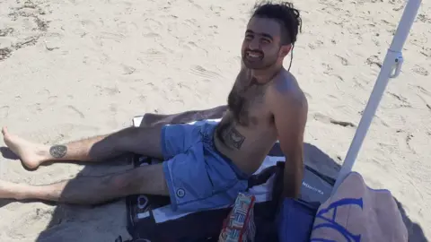 Un uomo sdraiato su una spiaggia sabbiosa che indossa pantaloncini, sorride alla telecamera