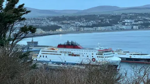 BBC The Manxman ferry, which is white, red and black, moored in Douglas Harbour in front of the Manannan fast craft, which is the same colours. It is dusk and the lights on Douglas seafront behind are beginning to glow.