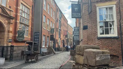 BBC News A cobbled street cluttered with historical objects including wagons, crates and lots of signage. The street is lined with Victorian warehouses and former shops and houses and has been made up to look like Victorian London, with signs advertising "artificial teeth" and "tinsmiths".