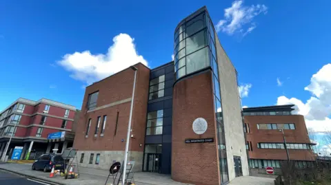 A photo of Hull Magistrates Court building. It has two floors and is a mix of brown brick and clear windows. There is a car parked in front and some roadwork signage and cones.