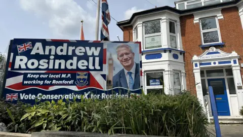 A large poster with a photo of Andrew Rosindell that says 'vote Conservative' stands in the front garden of a house that is used as the Tory party HQ in Romford. 