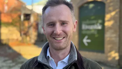 Charlie Homer, a man with short, light brown hair and a stubbly beard, is wearing a light blue shirt and a wax coat. He is standing outdoors in the centre of a town.