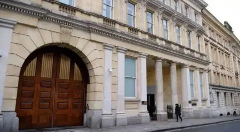 A grand white building with heavy set brown doors and pillars down the side of it.