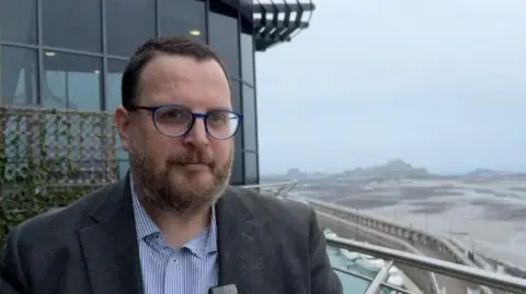 Chris Parker stands looking at the camera. He is wearing a suit and glasses. He has a beard and is standing on a balcony. 