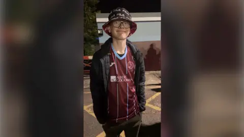 Family photo Harry is wearing a football shirt, jacket and tracksuit bottoms.  He is also wearing a bucket hat and glasses.  His hands are in his pockets. He is smiling at the camera.