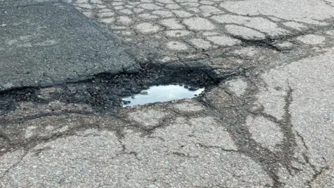 James Grant/BBC A large pothole filled with water with cracks in the road all around it.