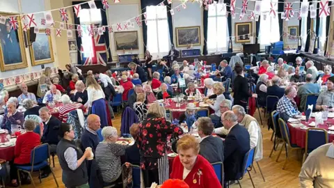 States of Jersey A picture of a number of people sat down at tables at a previous event. Tables are spread across the floor which is wood like. There is paintings on the wall around the side.