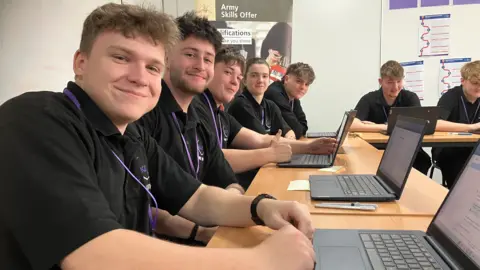 A row of schoolchildren wearing black polo shirts sitting at a desk with laptops and smiling at the camera.