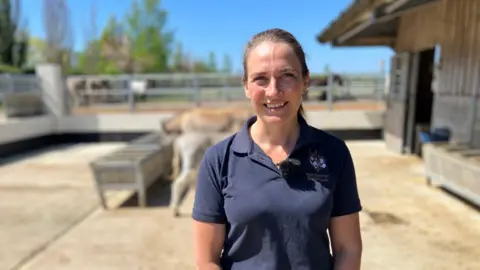 Jenna Goldby is standing outside at the sanctuary. Ms Goldby is looking at the camera and is smiling. She is wearing a navy blue polo top with the sanctuary logo embroidered on the shoulder. Two donkeys are standing behind her.