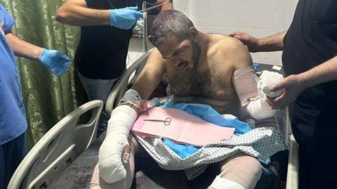 Amin Abed in a hospital bed with his arms and legs heavily bandaged (08/07/24)
