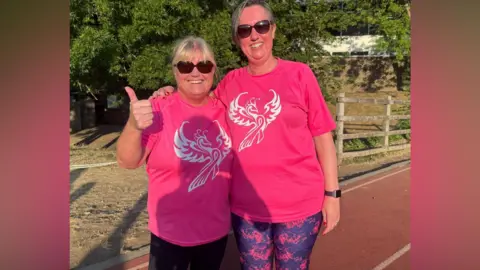 Jadzia Samuel/BBC Shirley Lowe and Leiann Bolton Clark standing side by side on a running track. Both are wearing pink t-shirts for their running team, Team Phoenix. Shirley has blonde hair and is doing a thumbs up. Leiann has shortish greying hair. Both are wearing sunglasses and smiling to camera.