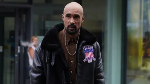 Ashley Warren crosses a street outside court wearing a brown shirt and a black leather jacket. He is looking past the camera.