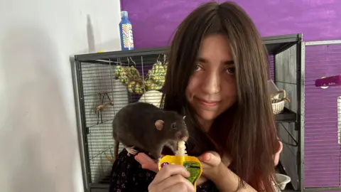 A young woman with long brown hair and a nose ring holds a rat, which is feeding from a pouch. She is standing in a room with white and purple walls, with a tall rat cage behind her.