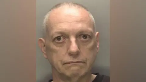 West Midlands Police A police custody photograph of a man with short grey hair and a gaunt face, wearing a dark top. He stares directly at the camera with a neutral expression. The West Midlands Police crest is visible in the bottom right corner of the image.
