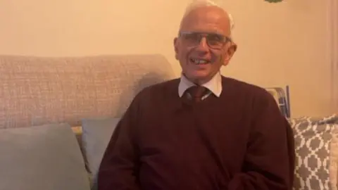 A man with short white hair wearing glasses is smiling and wearing a burgundy jumper with a burgundy tie and white shirt underneath. He is sat on a grey sofa with three grey pillows on it.