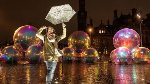 BD is LIT Several large inflatable bubbles featuring as part of an art installation in City Park - a public square in Bradford. A woman is stood in front of the installation wearing a rain jacket with her hood up, jeans and is holding an umbrella in the air.