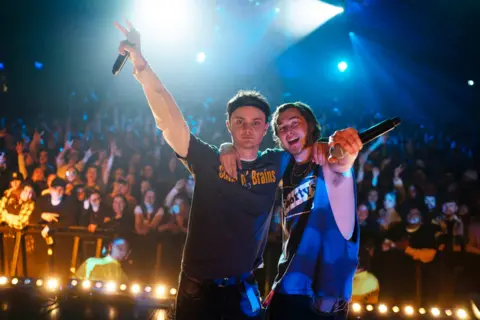 Studio Canal Two young male rappers stand on stage with one arm around each other, the other arm held aloft with microphone in hand. They have their backs to a large cheering crowd.