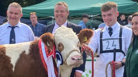 A brown and white cow is wearing red and white ribbons. A young man is holding his reins. He has light coloured hair and wearing a long white jacket. There are people standing about.