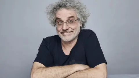 Aardman Animations Studio picture of Matt Perry - he has curly grey hair and glasses, smiling with arms crossed in front of him. He is wearing a blue t-shirt and has a small hoop earring in his left ear.