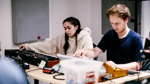 dBs Institute Two young people, one male and one female, practice operating a sound desk inside a room with light grey walls and soundproofing panels