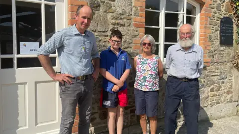 Charlie is wearing a blue T-shirt and shorts and is standing in front of a stone farmhouse his dad and grandparents and they are all smiling at the camera. 