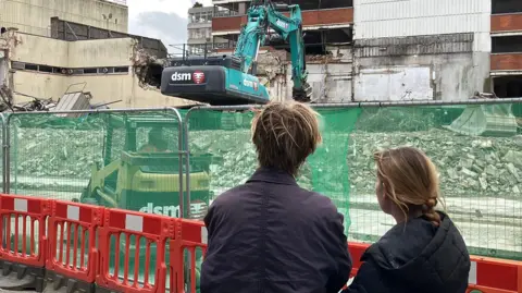 Two people are standing with their backs in front of the camera while a picture is being taken. They are both wearing black coats. They are standing in front of Anglia Square in Norwich which is currently in the process of being demolished. 