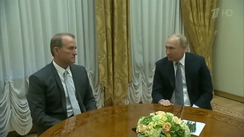 Channel One Russia Two men are sitting at a brown wooden table and looking at each other over a posy of yellow flowers. The man on his left is wearing a grey suit, white shirt and grey and white striped tie. On the right is the Russian President Vladimir Putin who is waring a dark grey suit, grey tie and white shirt and has a stack of papers in front of him 